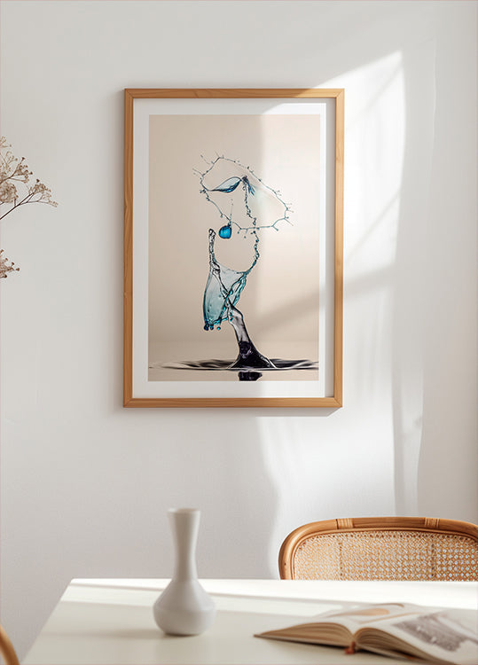 ein gerahmtes Kunstwerk eines in der Luft schwebenden Wassertropfens mit einem blau-weißen Farbschema. Das Kunstwerk ist an einer weißen Wand über einem Tisch mit einer weißen Vase und einem Buch ausgestellt.